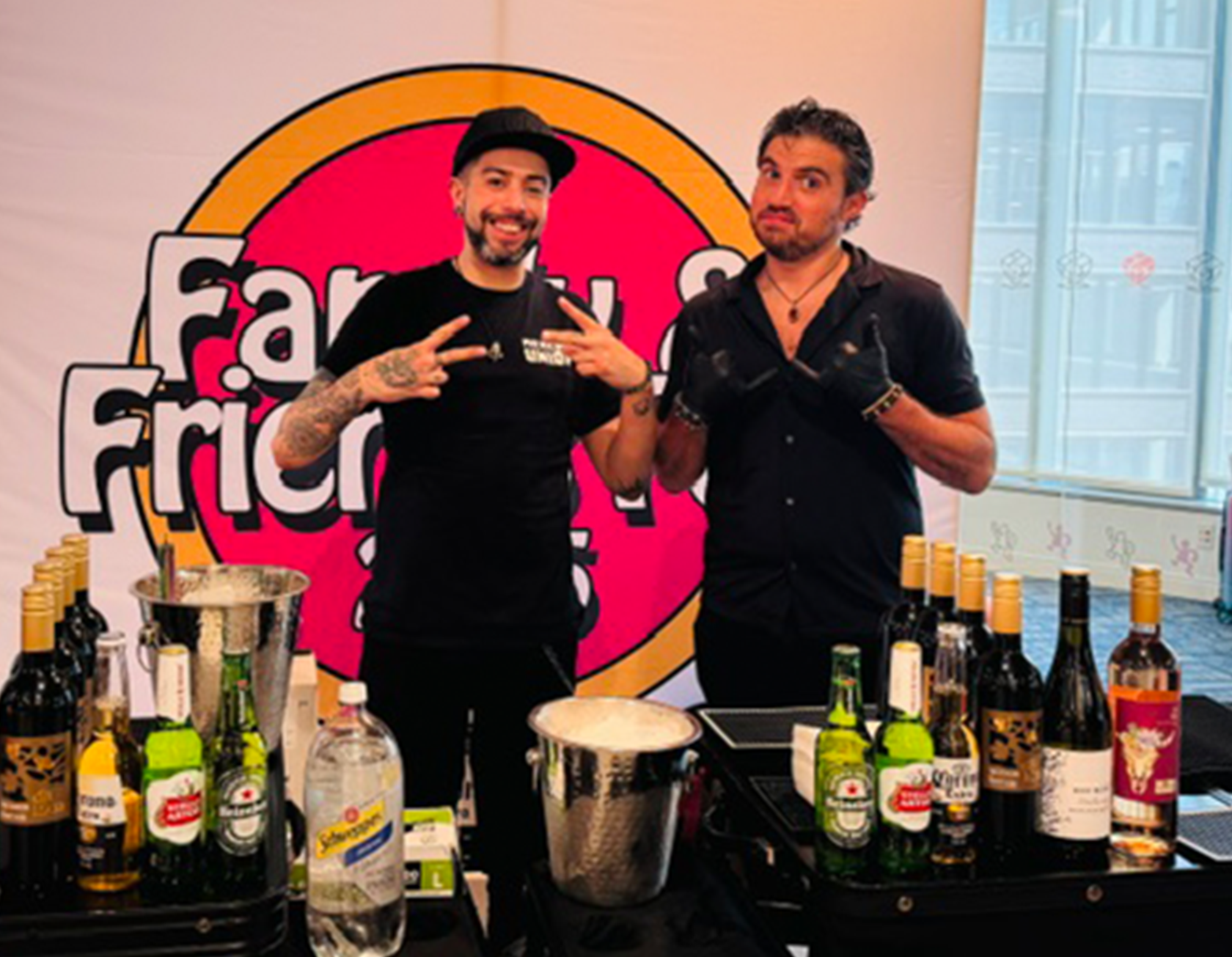 Two men standing behind a table with various bottles of alcohol and drink mixers, in front of a large sign that reads 'Farr Out Frier.' One man is wearing a black shirt and a cap, making a peace sign with his fingers, smiling. The other man, also in black, is pointing towards the first man, with a neutral expression. The background shows a window and parts of a building outside.