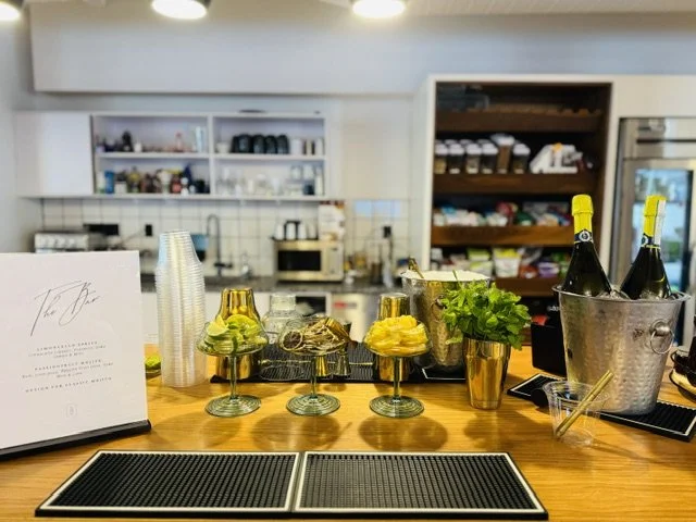 Bar counter with glasses of cocktails, bottles of champagne in a bucket, and fresh herbs, in a cafe or bar setting.