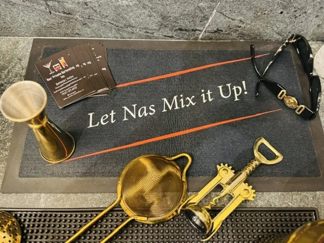 A black doormat with the phrase "Let Nas Mix it Up!" written in white, placed on a gray floor. On the mat, there is a gold-colored martini shaker, a gold-colored strainer, a gold-colored corkscrew, a pair of sunglasses, and a small cup. To the left s