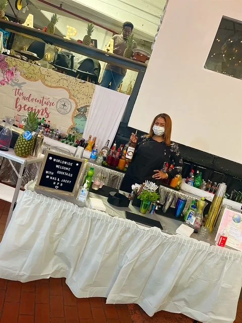 A woman wearing a face mask standing behind a bar with various bottles of alcohol. A sign on the table reads 'Worldwide Welcome with Cocktails I Was a Jacky N P B.' Behind her, there is a decorated wall with a sign that says 'The adventure begins' an