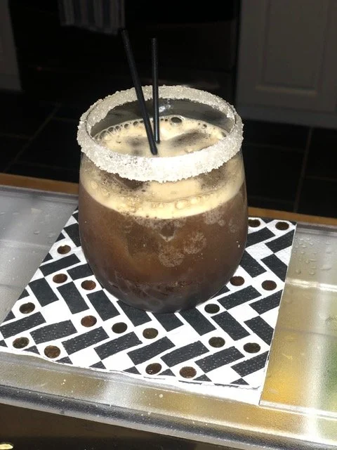 A glass of iced coffee with two black straws and a sugar rim, placed on a black and white patterned coaster.