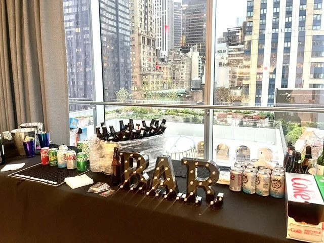Party table with bottled beverages, soda cans, and decorative lights spelling 'BAR' in front of large cityscape window.