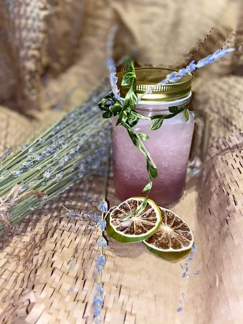 A mason jar with a gold lid containing a pink beverage, garnished with lavender sprigs, green leaves, and a decorative straw. Surrounding the jar are dried lemon slices, a lavender bundle, and a bundle of lavender tied with string, all on a woven sur