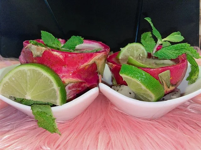 Two spicy watermelon margaritas garnished with lime wedges and mint leaves, served in hollowed-out watermelon rinds on white bowls, sitting on a pink furry table.