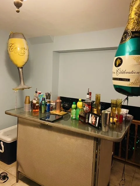 A party scene with two large champagne-shaped balloons, a bar counter with assorted drinks and cups, and a framed photograph, indicating a celebration.