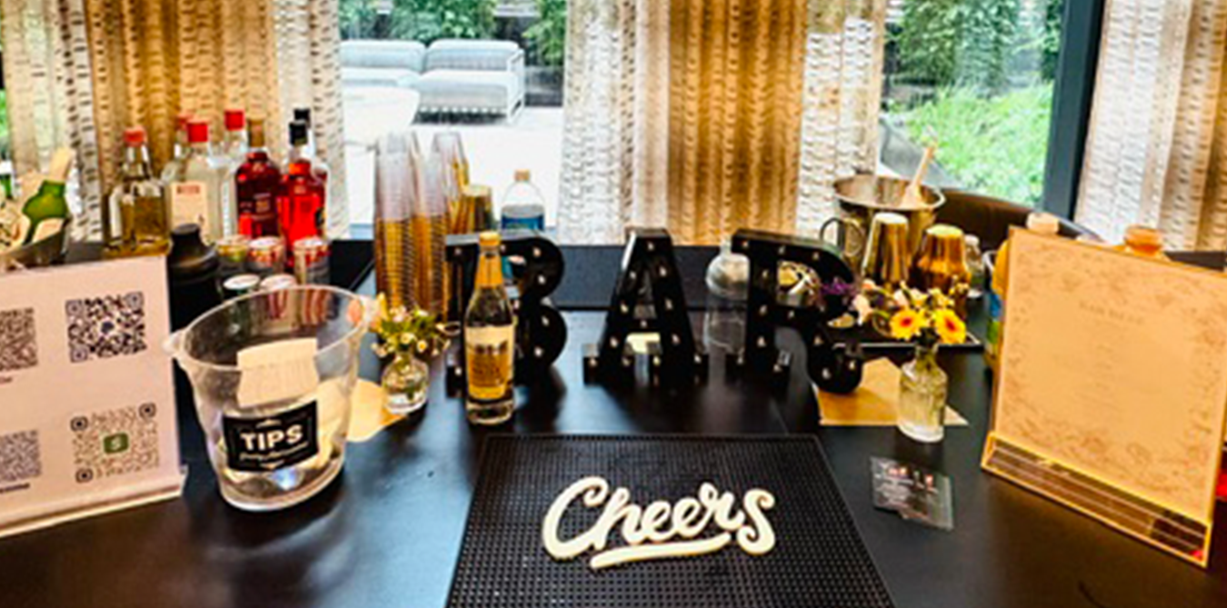 Party table with alcohol bottles, decorative letters spelling 'BAR', a vase with flowers, tips jar, and a 'Cheers' sign, in a room with curtains and a view outside.