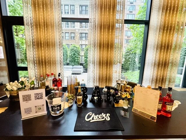 Indoor bar setup with bottles of alcohol, glasses, flowers, tips sign, a black mat with 'Cheers' written on it, and decor, with large windows and curtains showing a building and trees outside.