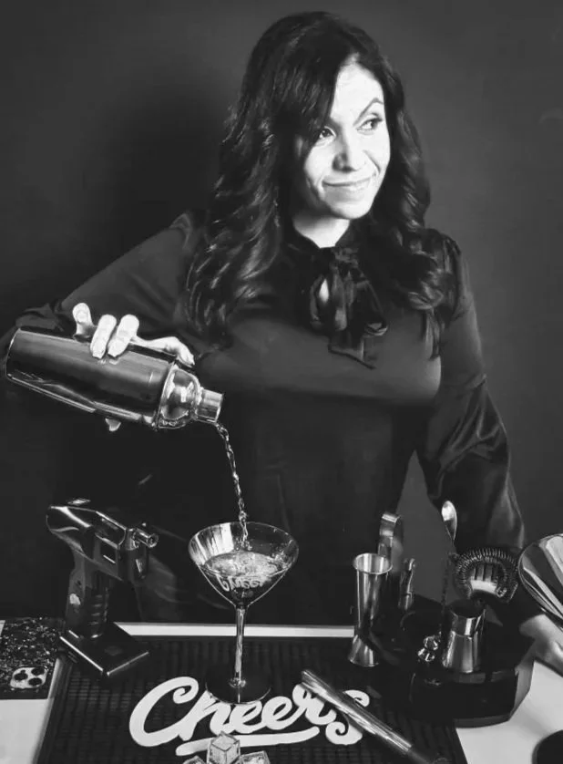 A woman with long dark hair pouring a cocktail from a shaker into a glass on a bar counter.
