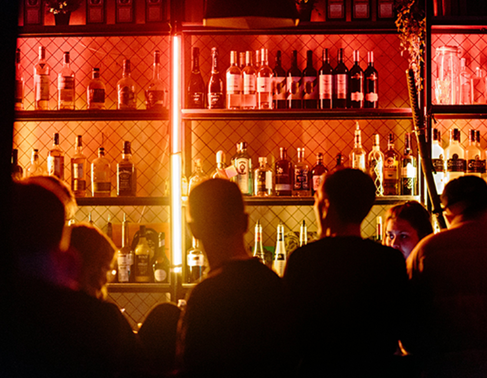 People gather in a dimly lit bar with a backlit display of various liquor bottles on shelves illuminated by red neon lights.