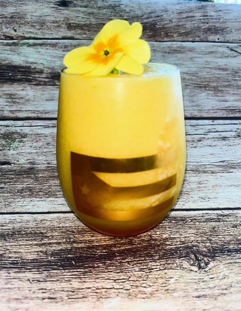 A yellow drink in a glass with a yellow flower garnishing the top, placed on a weathered wooden surface.