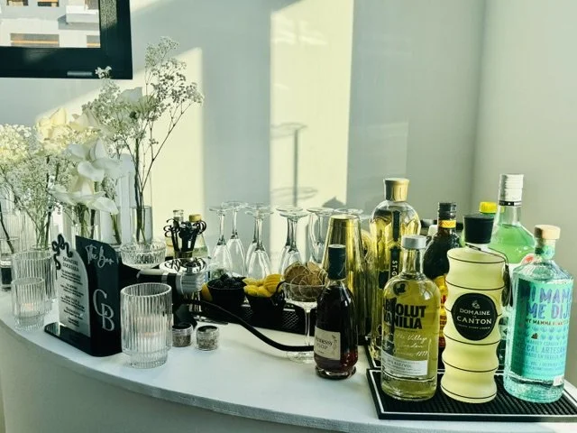 Bar setup on a white countertop with various bottles of alcohol, mixers, glasses, and decorative flowers.