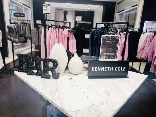 Display table in a retail store with clothing and decorative items. The table features white vases, a black sign with white text reading 'KENNETH COLE,' and decorative letters spelling 'PAR.' Behind the table are clothing racks with pink and blue gar