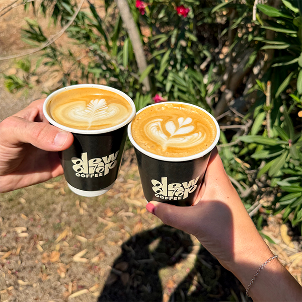 Two DewDrop Coffee branded cups of latte art coffee held up outdoors against a background of green foliage and pink flowers.