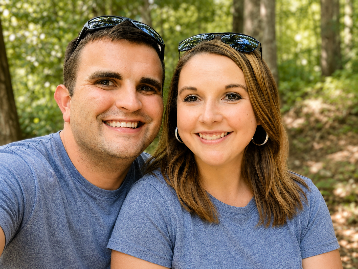 A smiling man and woman taking a selfie outdoors in a wooded area, both wearing blue shirts and sunglasses.