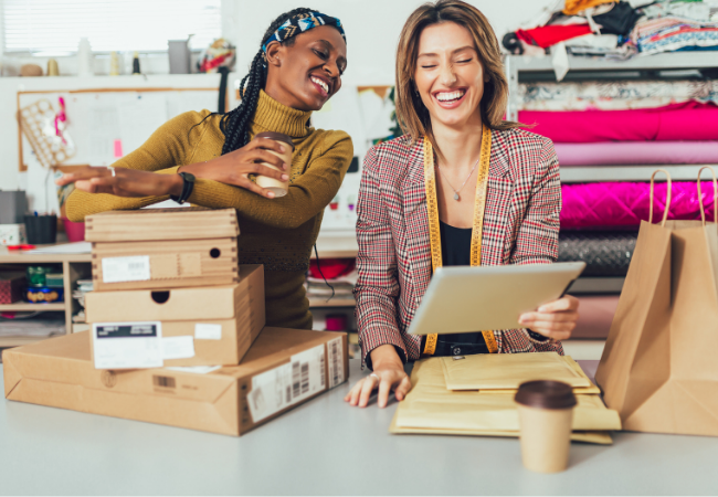 Two women smiling and engaging at a workspace with packages, fabric rolls, and a tablet.