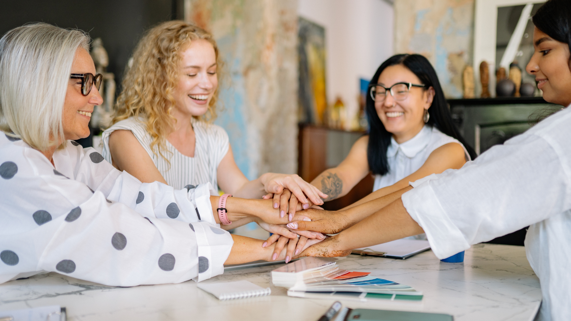 Group of diverse women with hands stacked together at a table, smiling during a meeting or team activity in a well-lit indoor space.