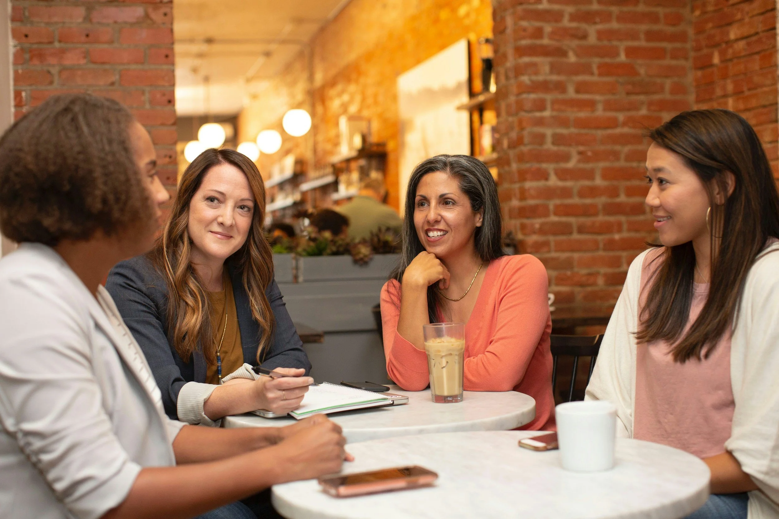 Four women sitting around a table in a restaurant, engaged in conversation. One woman is holding a phone, others are smiling or listening. The table has a glass of iced coffee, a drink, and a mug. The background features warm lighting, brick walls, and shelves with decorations.
