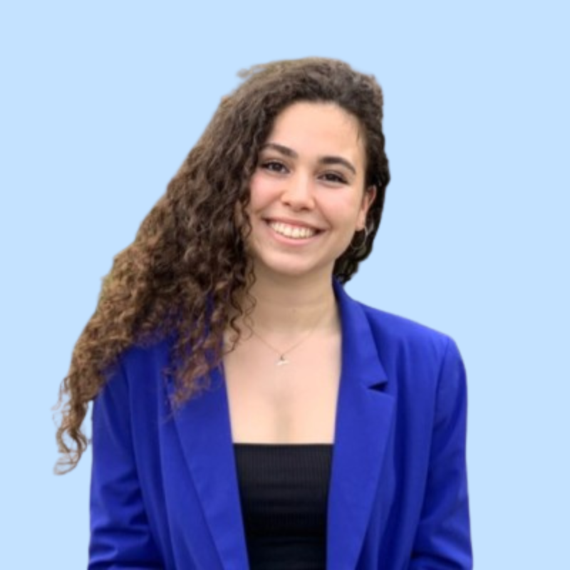 A young woman with curly brown hair smiling, wearing a royal blue blazer and a black top, against a light blue background.