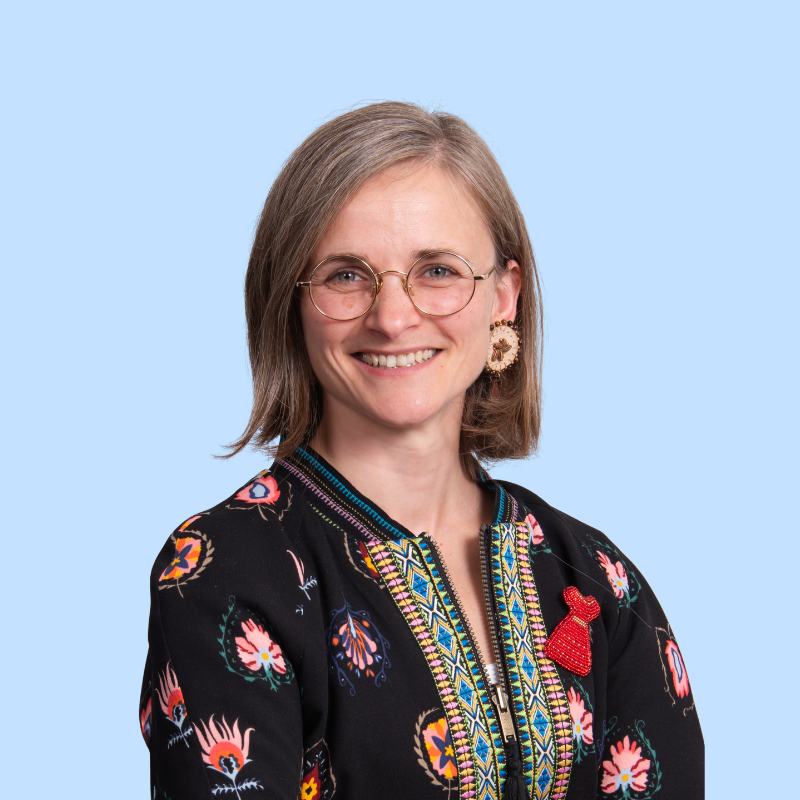 A woman with shoulder-length gray hair and glasses smiling, wearing a black embroidered jacket with colorful floral patterns, a large floral earring, and a red pin on her lapel, against a light blue background.