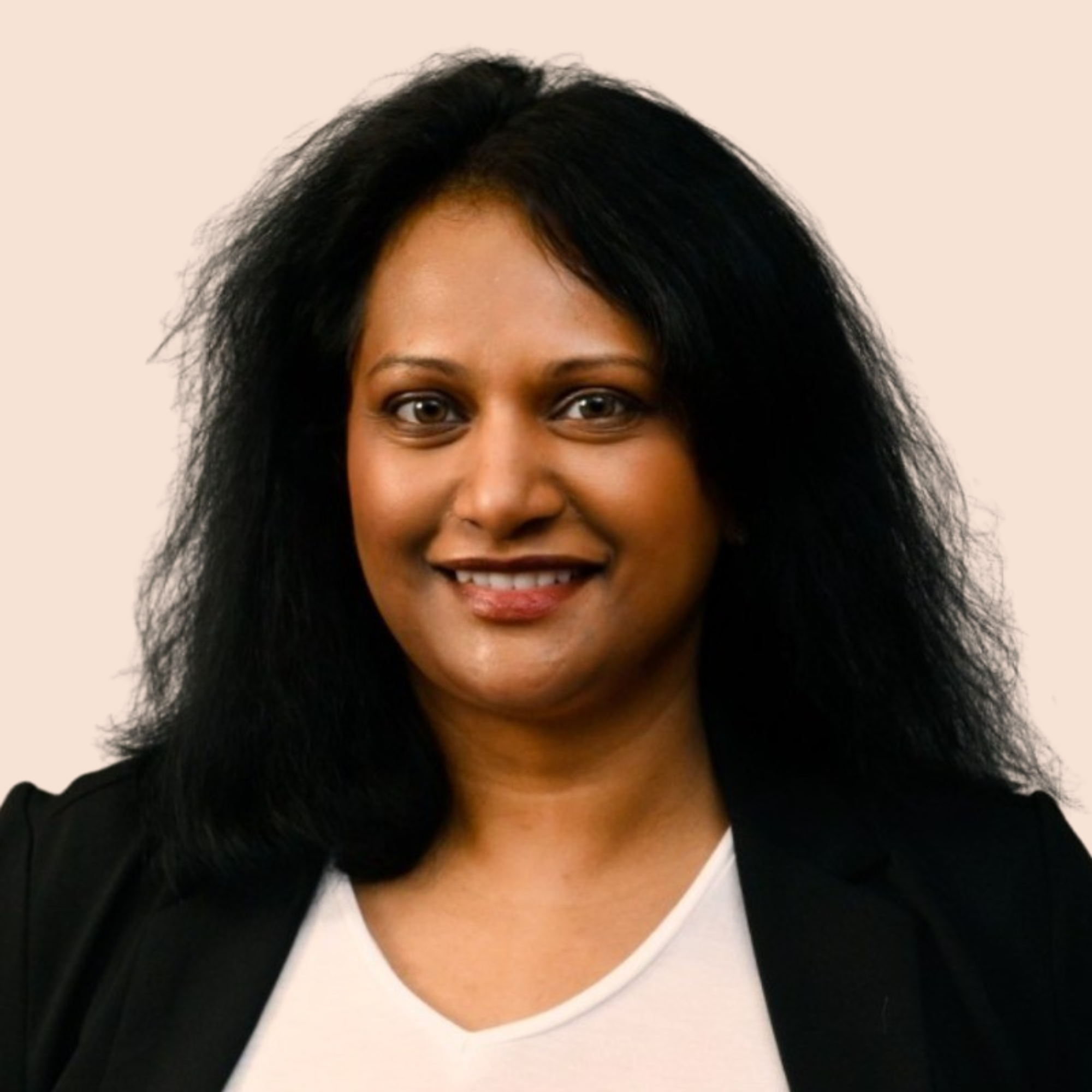 A woman with curly black hair, wearing a white shirt and a black blazer, smiling against a plain background.