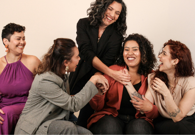 Five women sitting and standing together, smiling, and laughing during a friendly gathering.