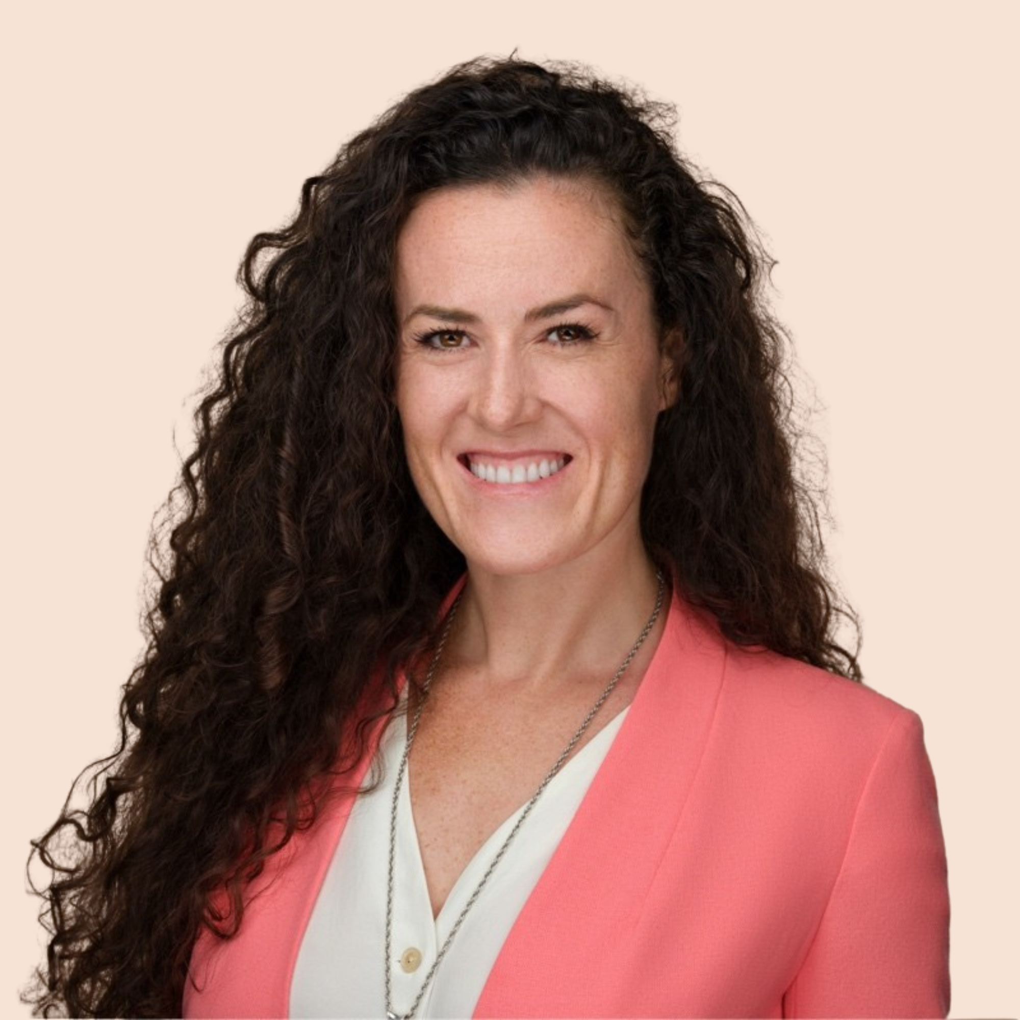 Close-up of a woman with long curly brown hair, smiling, wearing a coral blazer over a white blouse, and layered necklaces against a beige background.