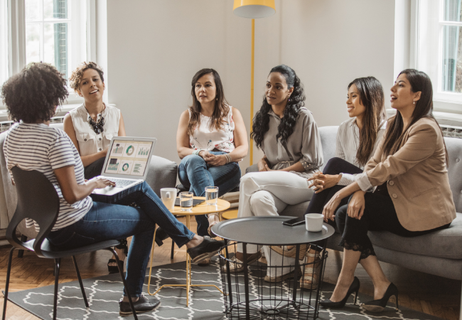 A group of six women sitting in a semi-circle in a well-lit living room, participating in a casual discussion or meeting, with one woman using a laptop, and various glasses of water and coffee cups on tables.