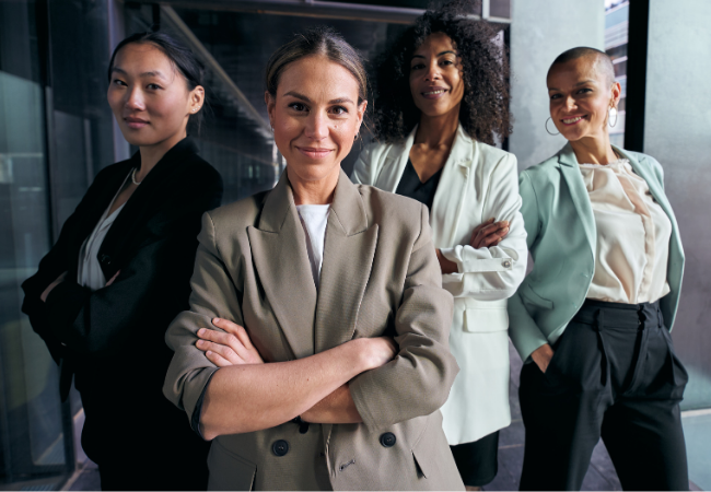 Four diverse women standing together in a modern office building, smiling and confident.