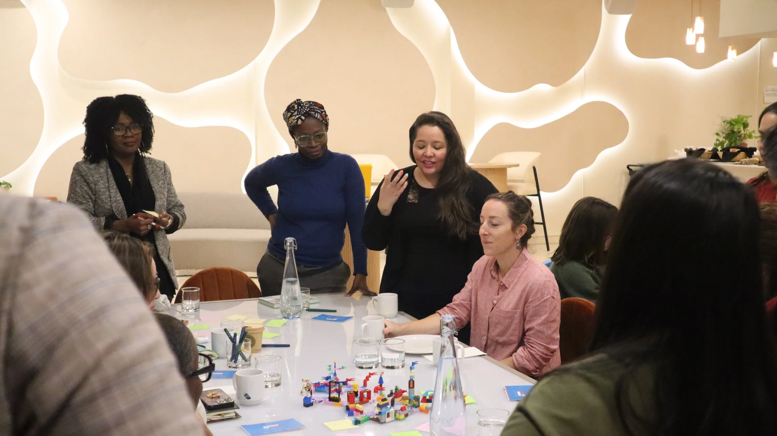 A group of women gathered around a table with colorful building blocks, notebooks, and drinks, engaged in discussion and activities in a modern room with decorative wall design.