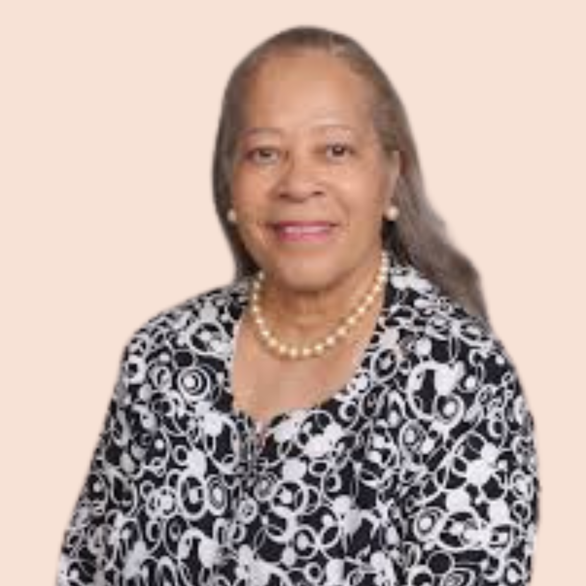 A woman with gray hair wearing a black and white patterned jacket, pearl earrings, a pearl necklace, and makeup smiling against a beige background.