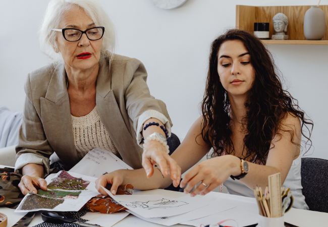 An elderly woman with glasses and a beige blazer assists a young woman with long, curly hair with a craft project at a table, surrounded by papers, fabric, and supplies.