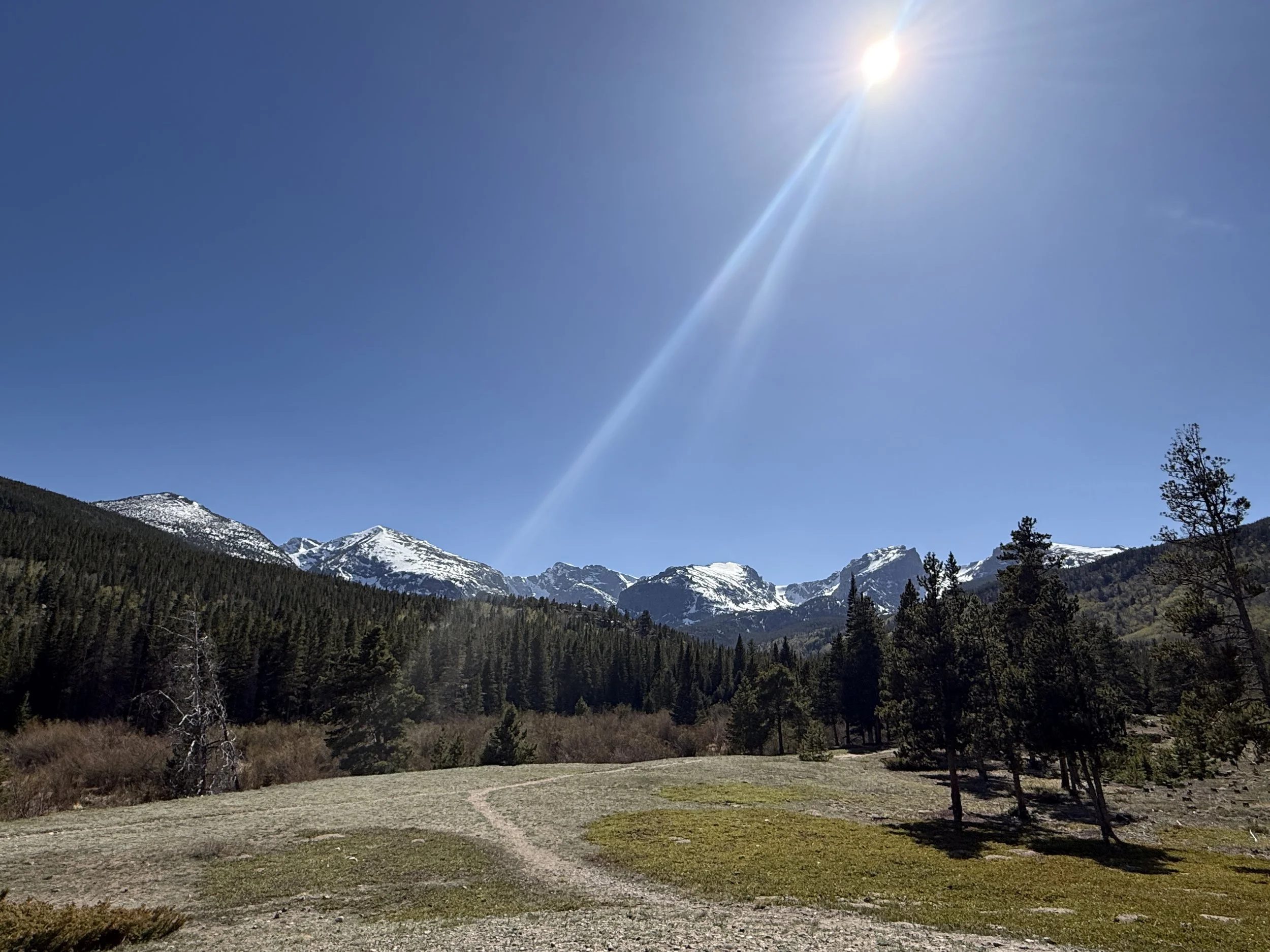 Mountains with snow caps in the background with trees and grass in the foreground and a sun with rays coming down in the sky near Denver, CO