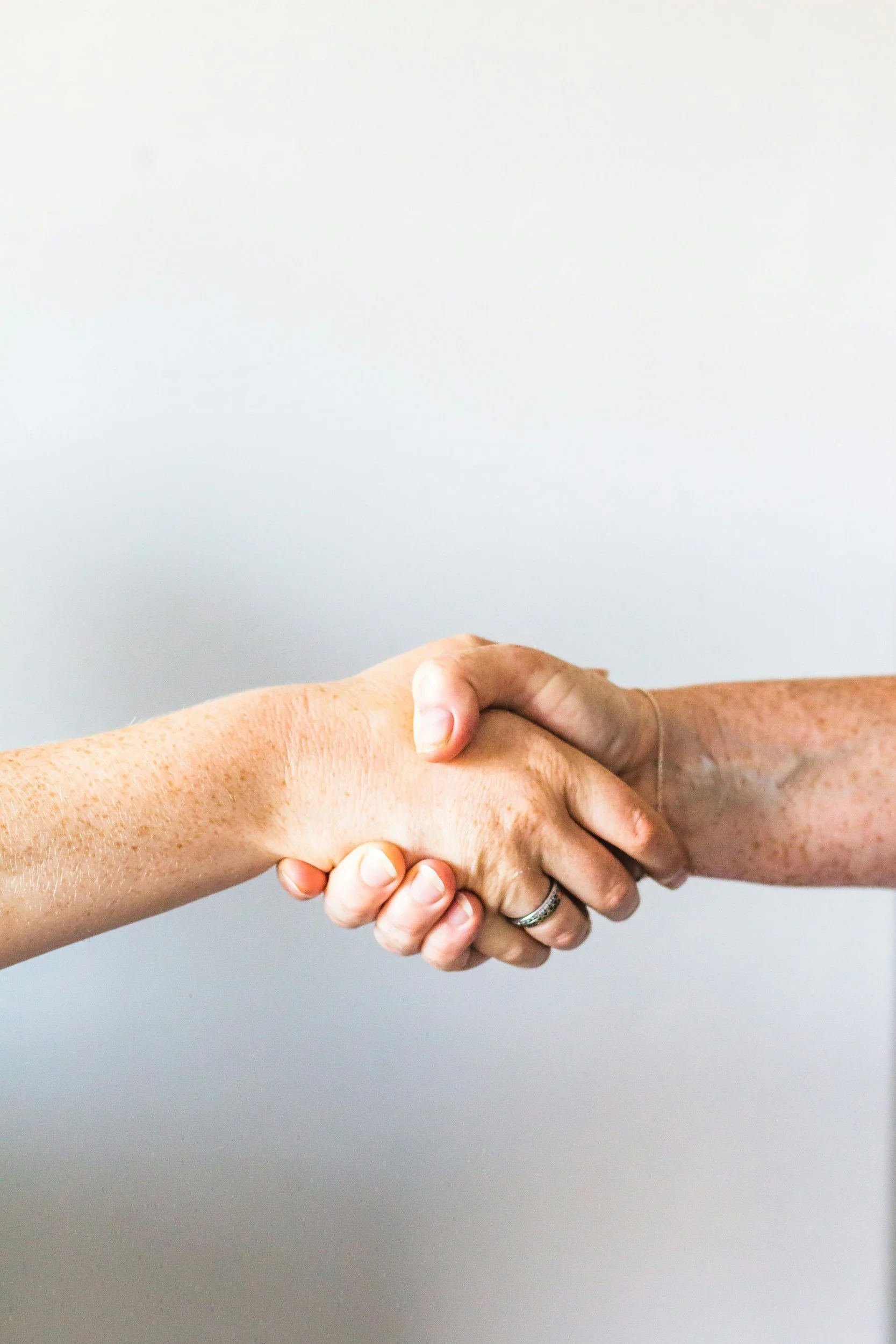 Two hands engaged in a handshake, with one hand appearing more aged and the other more youthful, against a plain background.