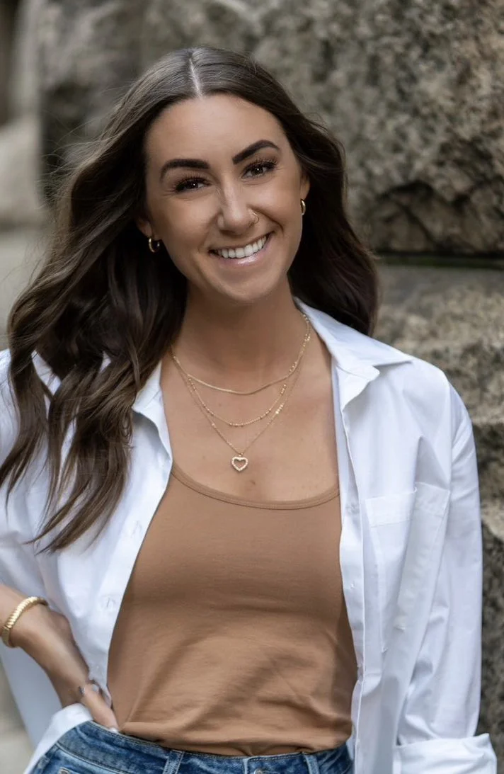 Smiling young woman with long brown hair, wearing a white shirt over a tan tank top and blue jeans, standing outdoors near rocks.