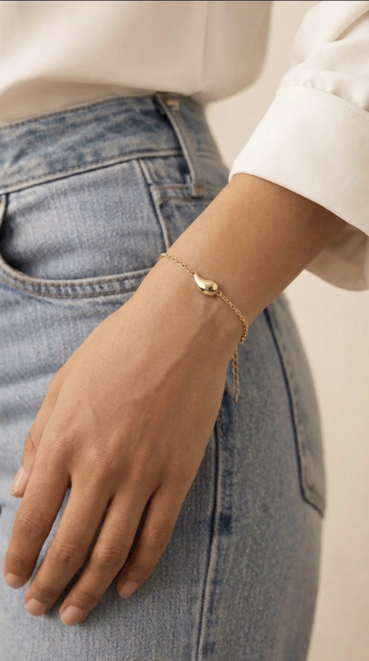 Close-up of a person's hand resting on their denim-jean-clad hip, wearing a delicate gold bracelet and a white shirt sleeve rolled up.