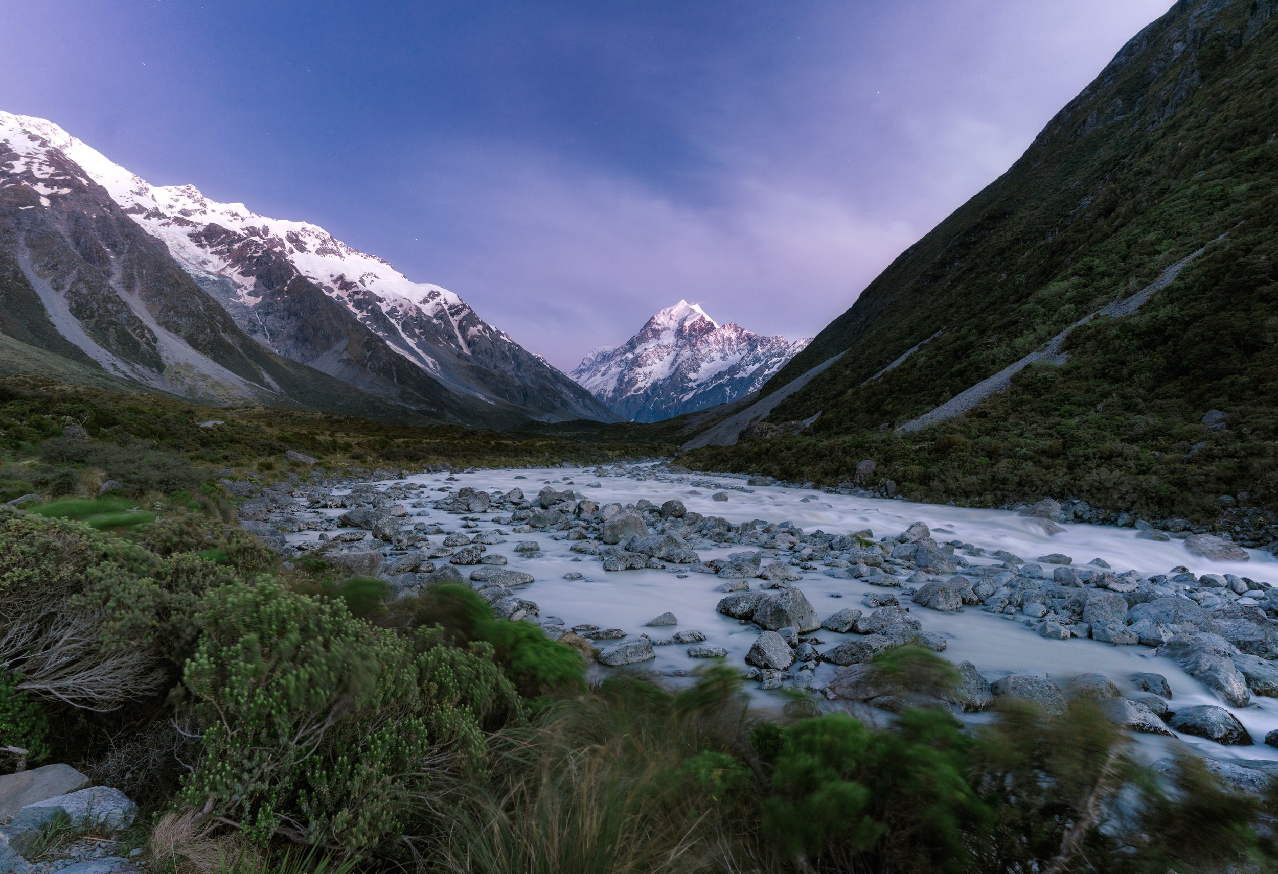 Scenic view of snow-capped mountains, a flowing river with rocks, and green vegetation under a clear evening sky.