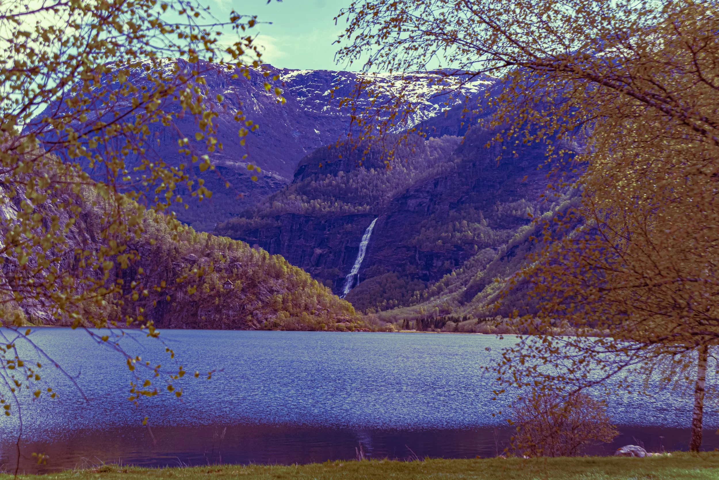 Scenic landscape featuring a lake surrounded by trees with yellow leaves, mountain cliffs with patches of snow, a waterfall, and a clear sky.