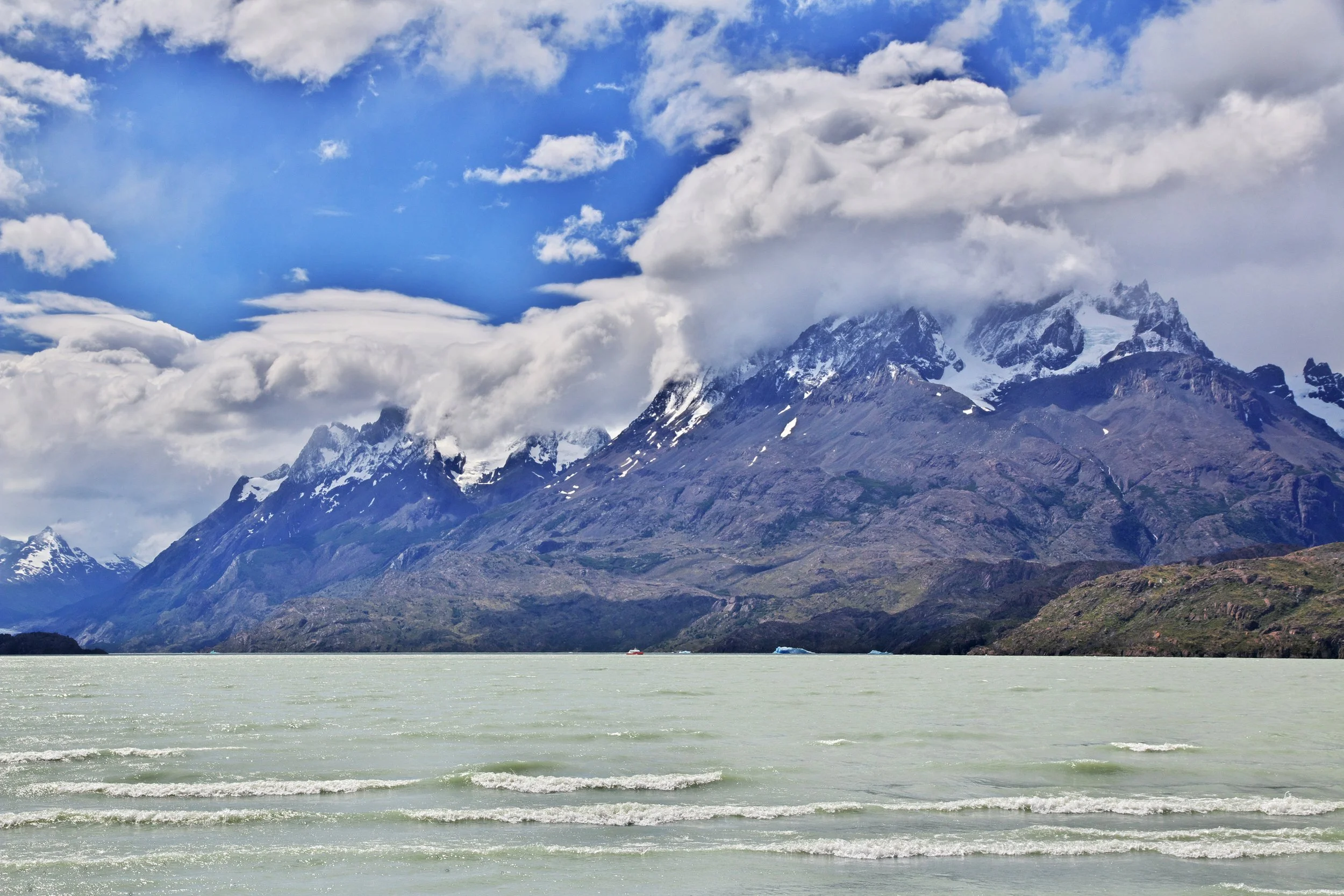 Scenic view of snow-capped mountains partially covered by clouds, with a large body of light green water in the foreground and a boat on the water.