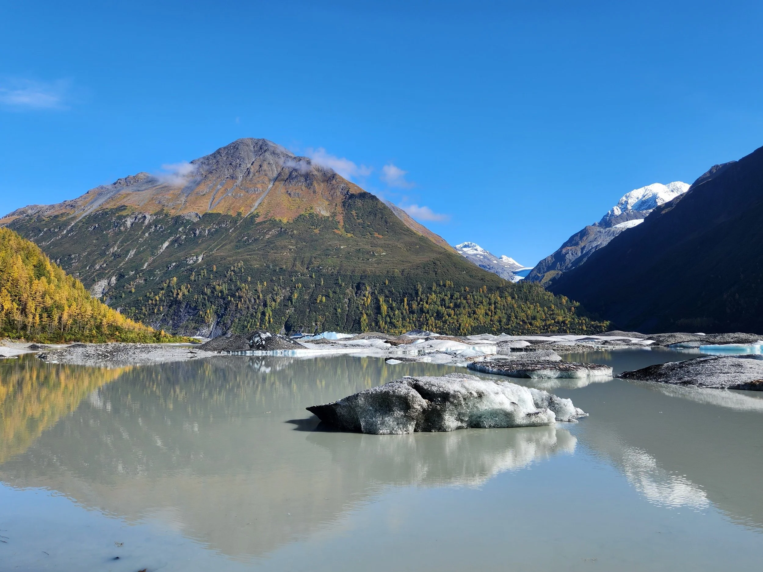 Scenic mountain landscape with green forested slopes, snowy peaks in the background, and a body of water with rocks in the foreground under a clear blue sky.