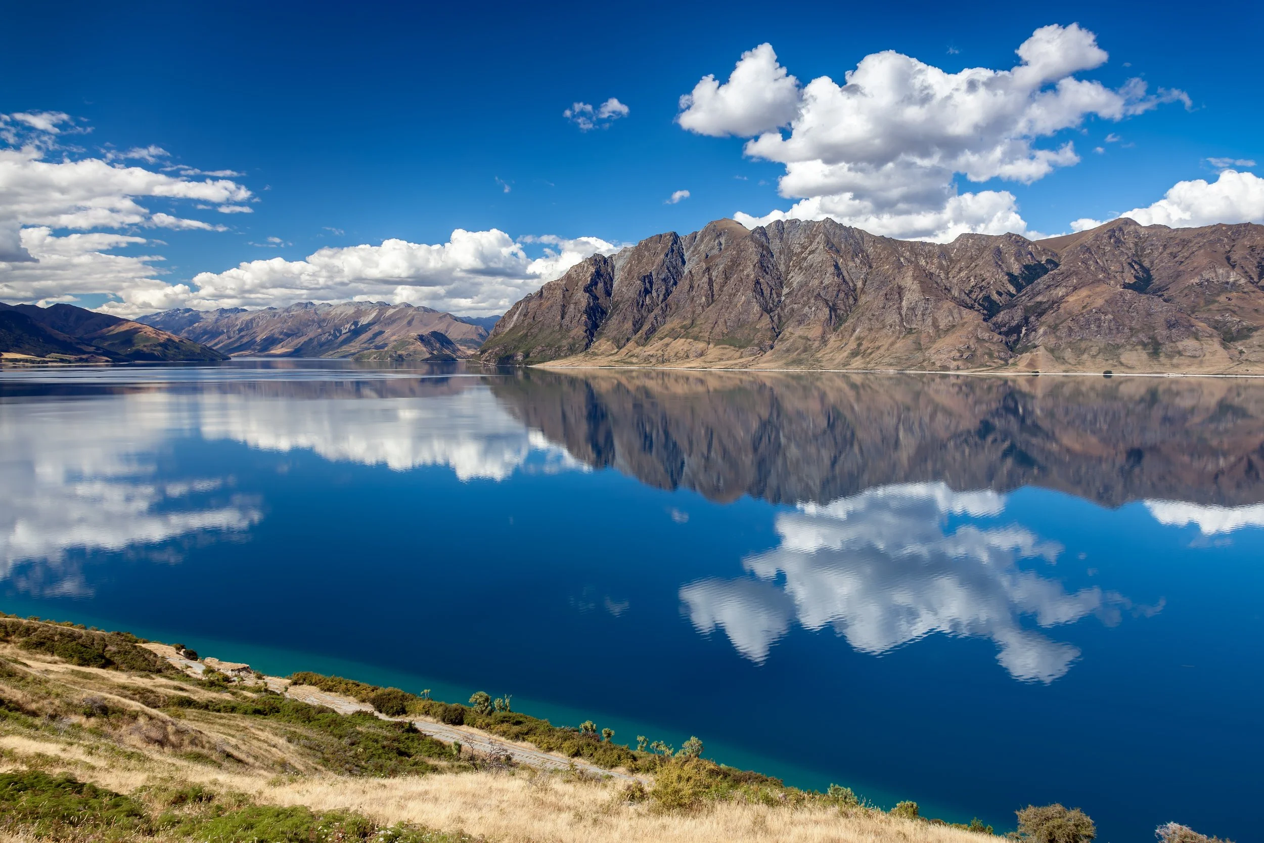 A large body of calm water reflecting the surrounding mountains and a partly cloudy blue sky.