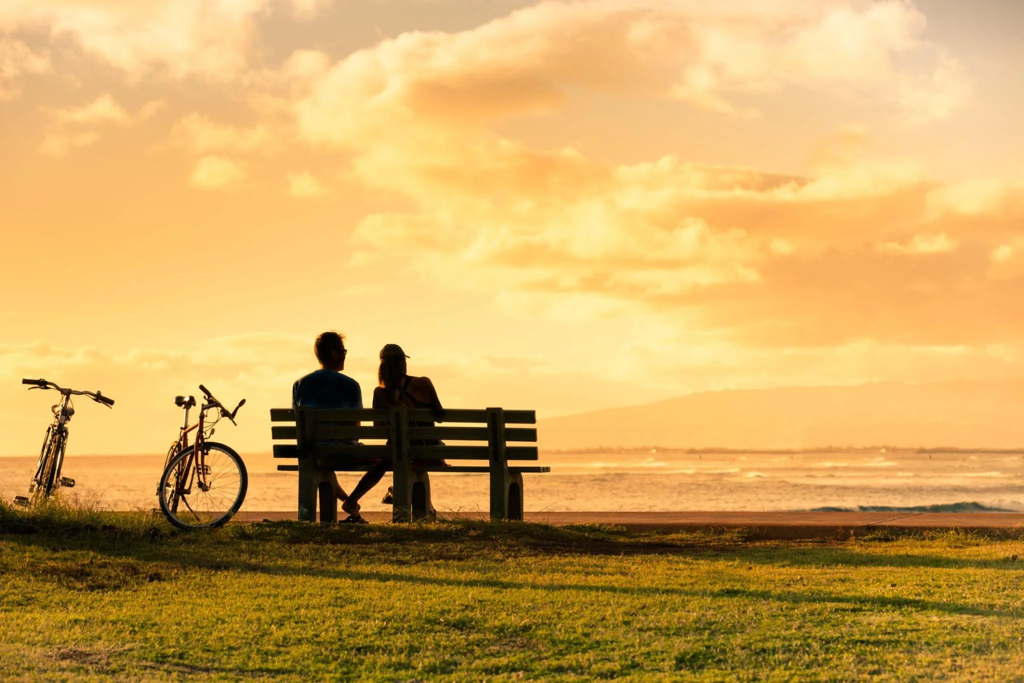 Two people sitting on a park bench at the beach during sunset, with bicycles parked nearby and the ocean in the background.