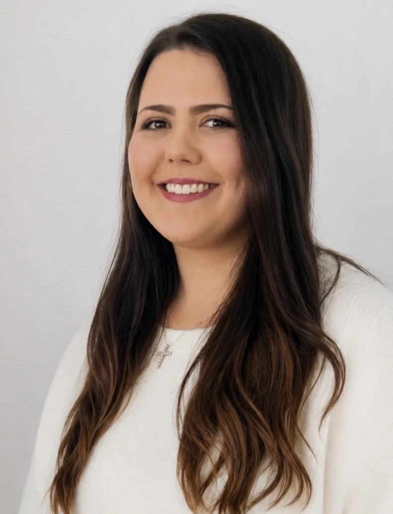 A young woman with long brown hair, smiling and wearing a white top and a necklace with a cross pendant, against a plain light background.