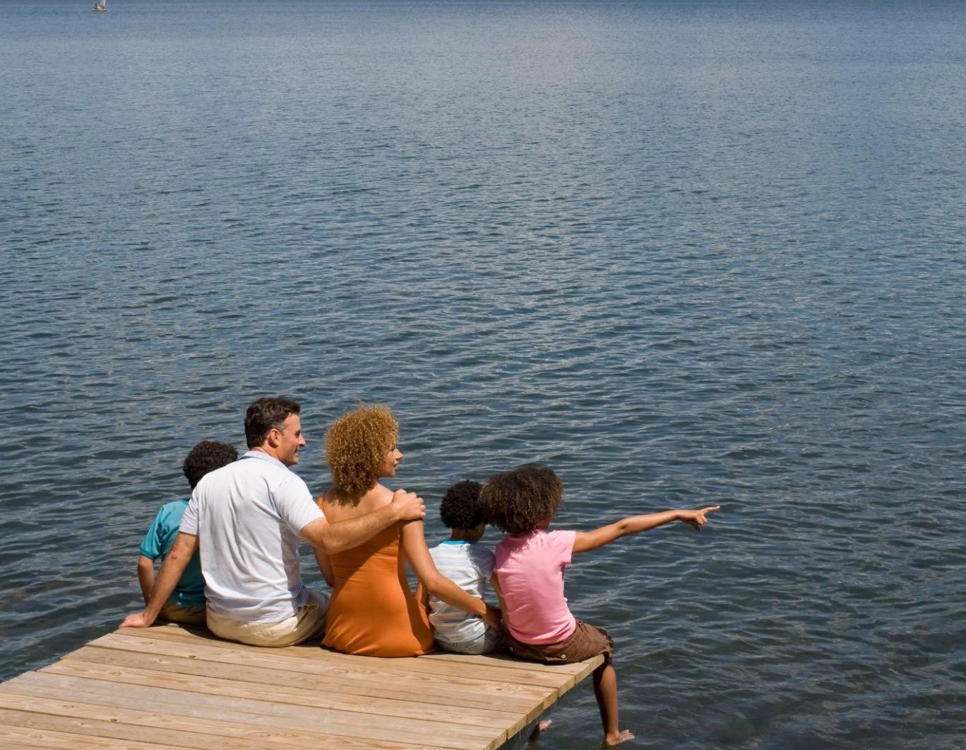Group of five people, including two adults and three children, sitting on a wooden dock at the edge of a body of water. One girl is pointing towards the water.