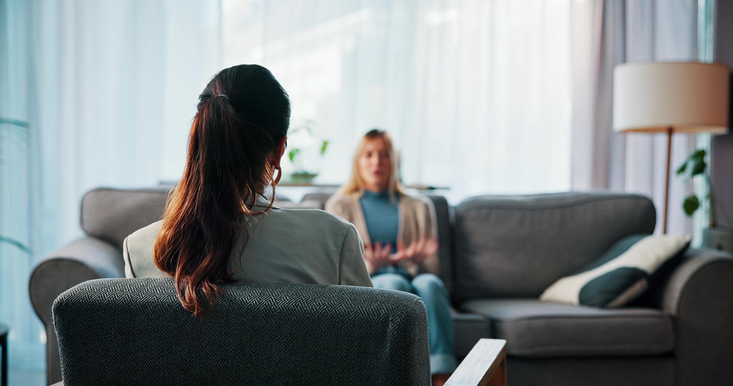 Two women having a conversation in a living room. One woman with dark hair in a ponytail is sitting in the foreground with her back to the camera, while the other woman, with blonde hair, is sitting in the background facing her and speaking.