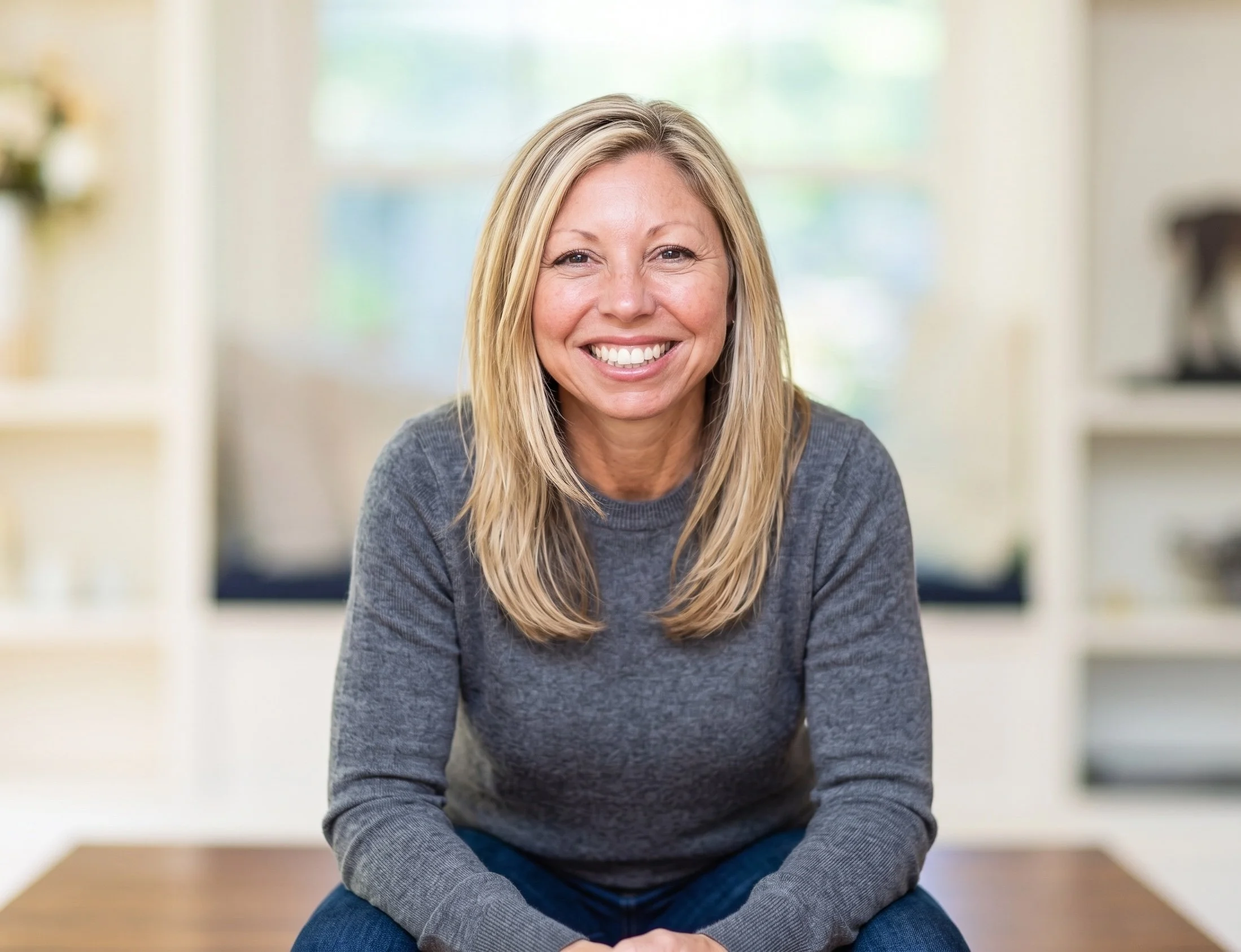 A smiling woman with long blonde hair and freckles wearing a gray sweater, sitting in a bright room with large windows and light-colored walls.