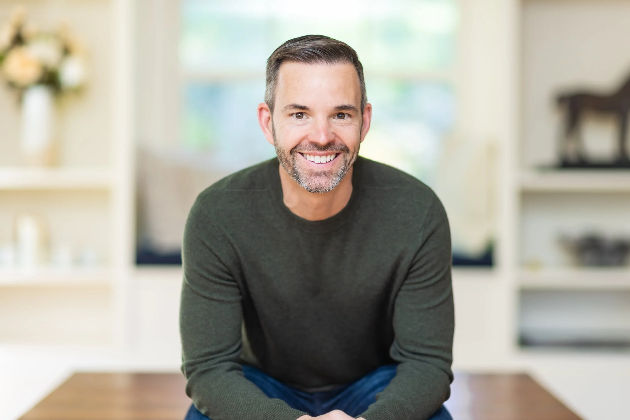 Smiling man with short, dark hair and a beard, sitting indoors with blurred background, wearing a dark green sweater.