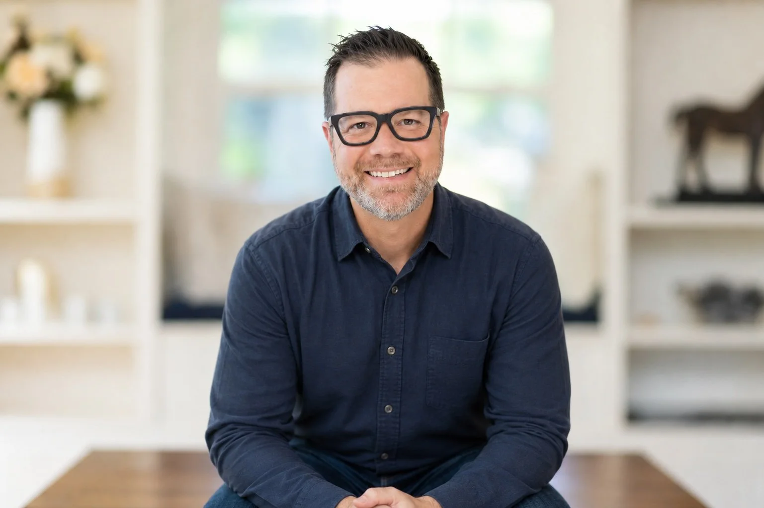 A middle-aged man with dark hair, glasses, and a beard smiling at the camera. He is wearing a navy blue button-up shirt and is sitting indoors with a blurred background of a room with shelves and a window.