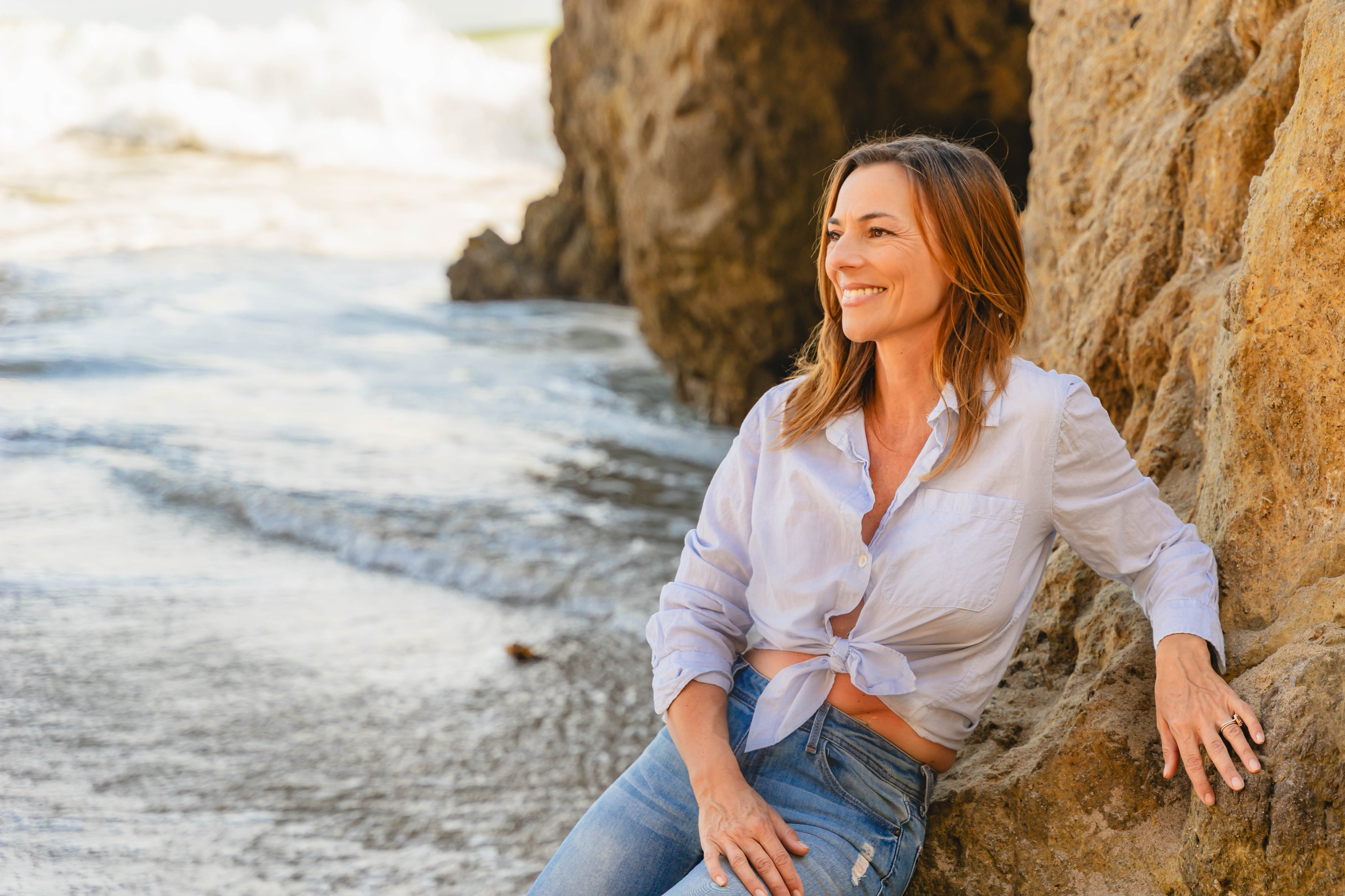 A woman with shoulder-length brown hair smiling while leaning against a large rock near the beach.
