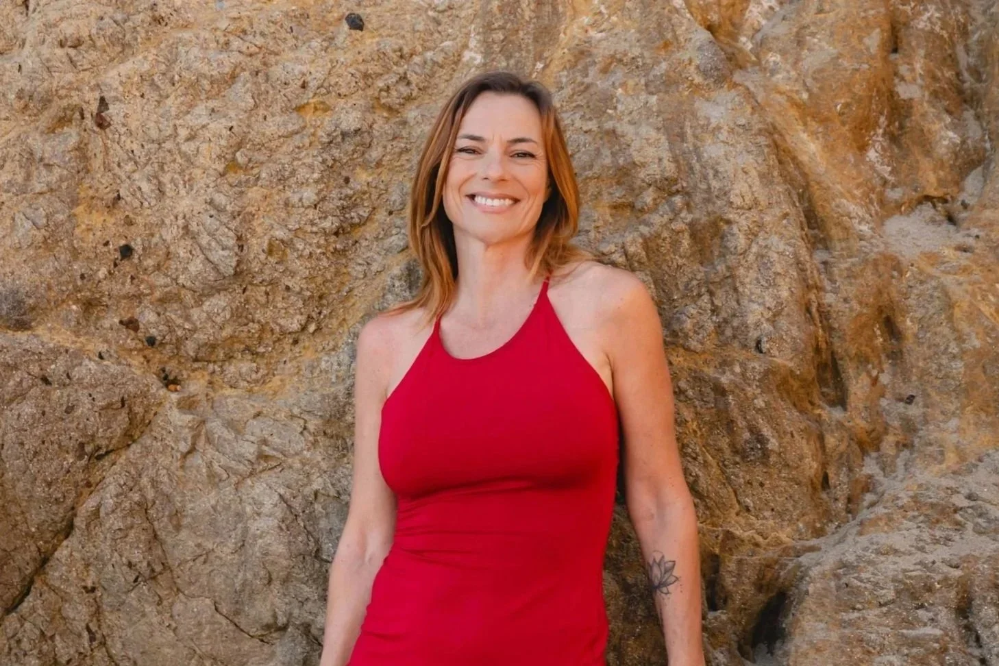 A woman with shoulder-length auburn hair, wearing a red sleeveless top, standing in front of a rocky background, smiling at the camera.