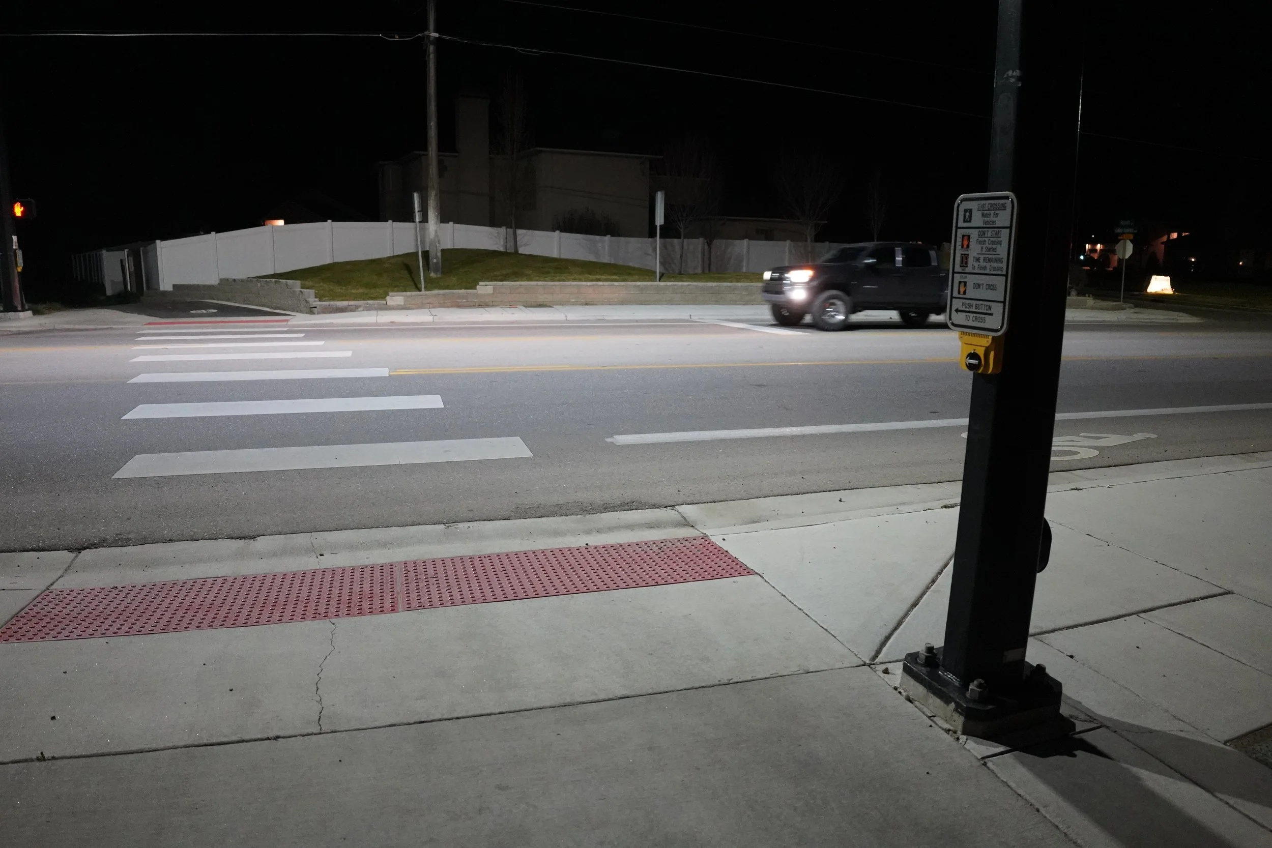 Nighttime street scene showing crosswalk, traffic signal, and a car passing by. A sidewalk with tactile paving is visible in the foreground. A white fence surrounds a small grassy area and some trees in the background. A street crossing button and traffic sign are mounted on a black pole to the right.
